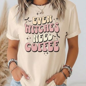 以下が含まれることがあります： クリーム色の半袖Tシャツで、レトロな波状のフォントで「EVEN WITCHES NEED COFFEE」というテキストがプリントされています。グラフィックには、前面の文字を囲むように小さな黒いコウモリのシルエットと星のアクセントが描かれています。