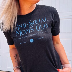 May include: Dark grey t-shirt with the text "ANTI-SOCIAL MOM'S CLUB" in light blue. The shirt also features the phrase "SOCIAL BATTERY: PERMANENTLY LOW" and a geometric design. The shirt is a crew neck.