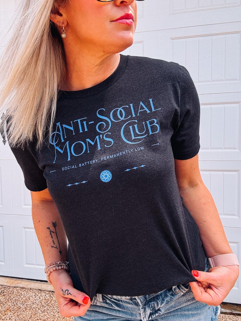 May include: Dark grey t-shirt with the text "ANTI-SOCIAL MOM'S CLUB" in light blue. The shirt also features the phrase "SOCIAL BATTERY: PERMANENTLY LOW" and a geometric design. The shirt is a crew neck.