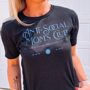 May include: Dark grey t-shirt with the text "ANTI-SOCIAL MOM'S CLUB" in light blue. The shirt also features the text "SOCIAL BATTERY: PERMANENTLY LOW" and a small graphic. The shirt is short-sleeved.