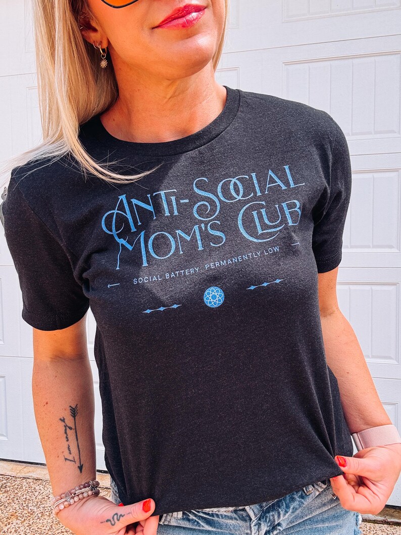 May include: Dark grey t-shirt with the text "ANTI-SOCIAL MOM'S CLUB" in light blue. The shirt also features the text "SOCIAL BATTERY: PERMANENTLY LOW" and a small graphic. The shirt is short-sleeved.