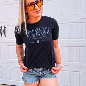 May include: A dark grey t-shirt with the blue text "ANTI-SOCIAL MOM'S CLUB" and "SOCIAL BATTERY: PERMANENTLY LOW". The person is wearing denim shorts and orange-tinted sunglasses. The image is taken in front of a white garage door.