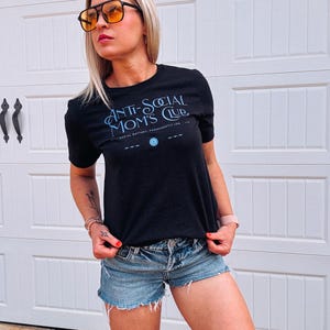 May include: Black crew neck t-shirt with the text "ANTI-SOCIAL MOMS CLUB" in light blue script. The shirt also features a small graphic and the text "SOCIAL BATTERY: PERMANENTLY LOW". The model is wearing denim shorts and orange-tinted sunglasses.