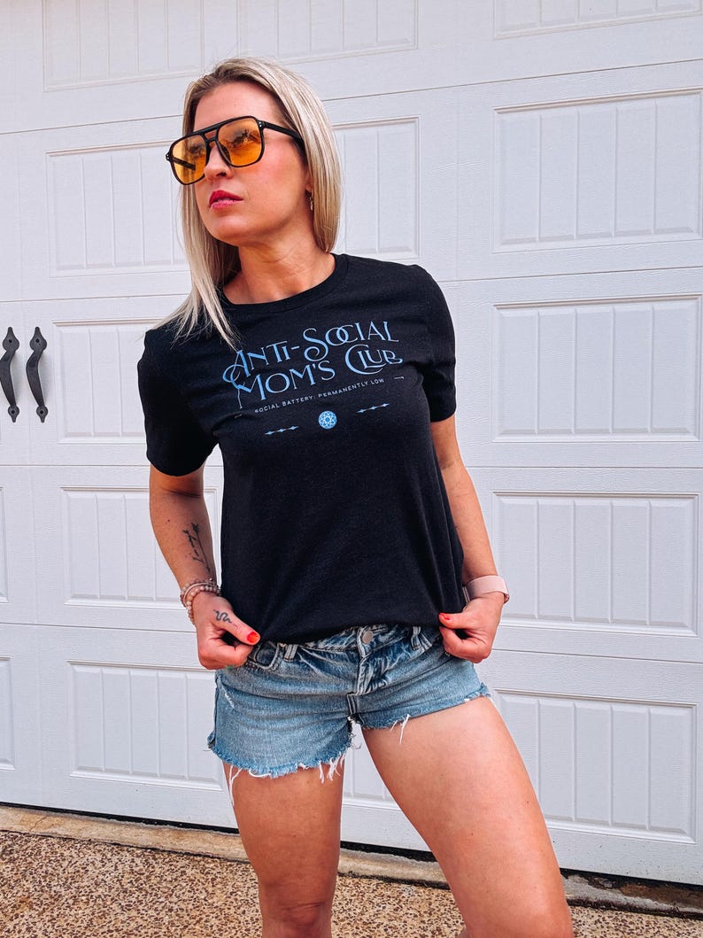 May include: Black crew neck t-shirt with the text "ANTI-SOCIAL MOMS CLUB" in light blue script. The shirt also features a small graphic and the text "SOCIAL BATTERY: PERMANENTLY LOW". The model is wearing denim shorts and orange-tinted sunglasses.