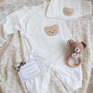 Pu&ograve; includere: Un completo per neonato bianco con un motivo a orsetto, composto da una tutina e un cappello abbinato. Un sonaglio a forma di orsetto all'uncinetto e un biglietto regalo con la scritta "Welcome, Little One! You are a TREASURE!" completano il set.