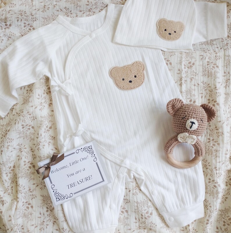 Pu&ograve; includere: Un completo per neonato bianco con un motivo a orsetto, composto da una tutina e un cappello abbinato. Un sonaglio a forma di orsetto all'uncinetto e un biglietto regalo con la scritta "Welcome, Little One! You are a TREASURE!" completano il set.