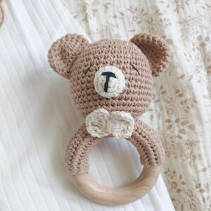 Pu&ograve; includere: Un sonaglio per bambini fatto a mano all'uncinetto a forma di orsetto. L'orsetto &egrave; marrone chiaro con un muso bianco e un naso nero. Ha un anello di legno per il bambino. Il sonaglio &egrave; appoggiato su una superficie bianca.
