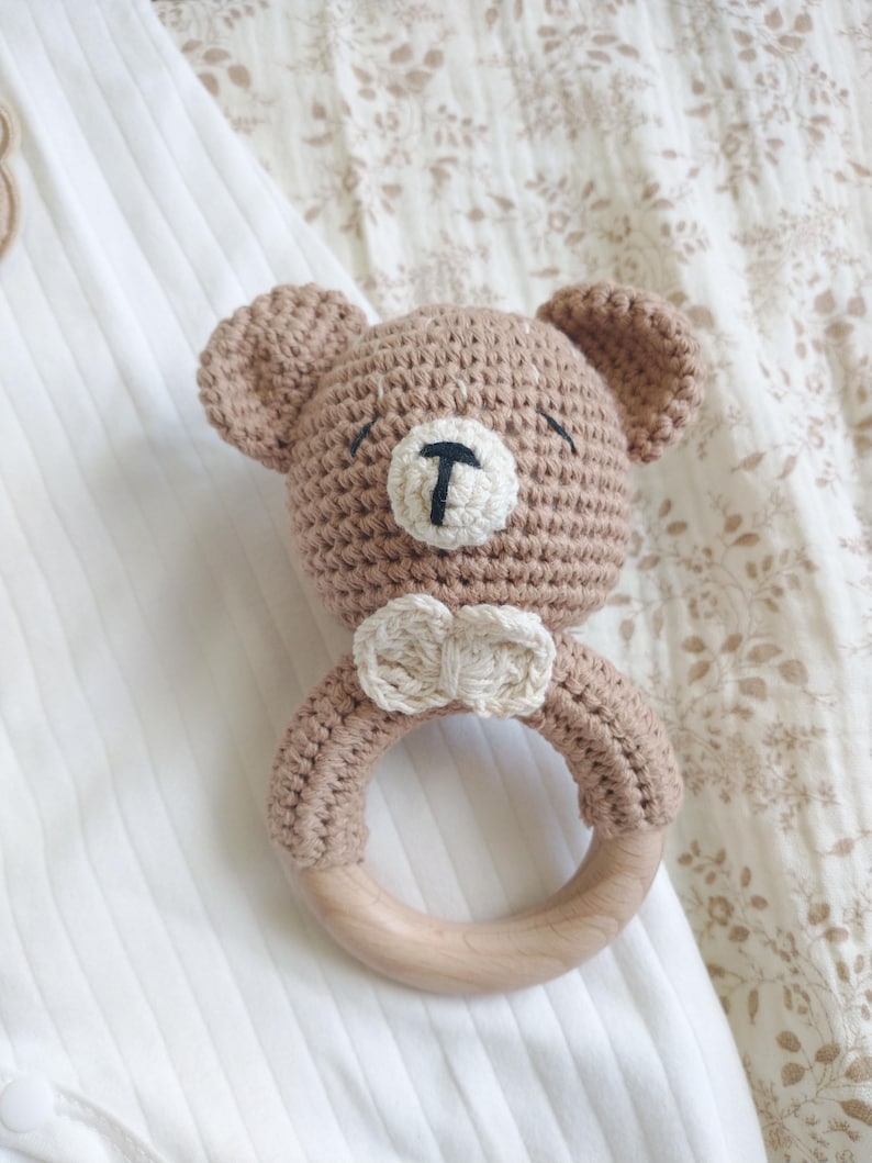 Pu&ograve; includere: Un sonaglio per bambini fatto a mano all'uncinetto a forma di orsetto. L'orsetto &egrave; marrone chiaro con un muso bianco e un naso nero. Ha un anello di legno per il bambino. Il sonaglio &egrave; appoggiato su una superficie bianca.