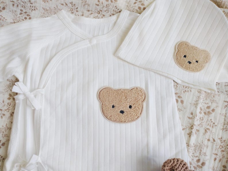 Pu&ograve; includere: Un completo per neonati bianco a coste, composto da un body avvolgente e un berretto abbinato. Ogni pezzo presenta un'applicazione a forma di orsetto marrone chiaro. Il set &egrave; realizzato in materiale morbido, per il comfort.