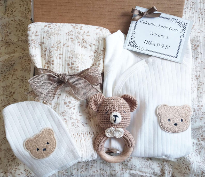 Pu&ograve; includere: Un set regalo per bambini in una scatola di cartone, tra cui un body bianco con una toppa a forma di orso, un cappello abbinato, una coperta a fantasia floreale e un sonaglio a forma di orso all'uncinetto. Una carta dice: "Benvenuto, Piccolo! Sei un TESORO!"