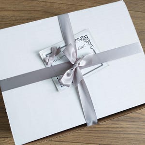 Pu&ograve; includere: Una scatola regalo rettangolare bianca legata con un nastro e un fiocco di raso grigio. Un piccolo biglietto con il testo "Welcome One!" &egrave; infilato sotto il nastro. La scatola &egrave; chiusa e pronta per essere regalata.