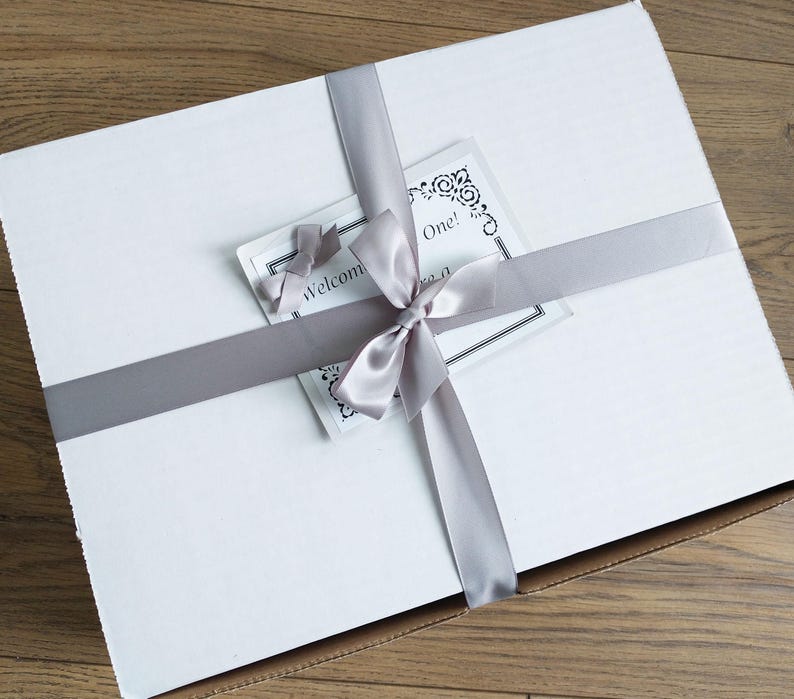 Pu&ograve; includere: Una scatola regalo rettangolare bianca legata con un nastro e un fiocco di raso grigio. Un piccolo biglietto con il testo "Welcome One!" &egrave; infilato sotto il nastro. La scatola &egrave; chiusa e pronta per essere regalata.
