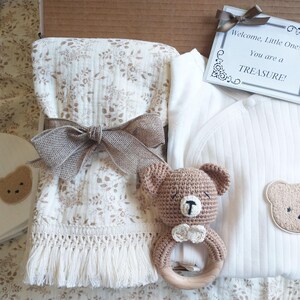 Pu&ograve; includere: Un set regalo per neonati con un body bianco con un'applicazione a forma di orsetto, una coperta con stampa floreale, un sonaglio a forma di orsetto e un biglietto con la scritta "Welcome, Little One! You are a TREASURE!". Il set &egrave; legato con un nastro marrone.