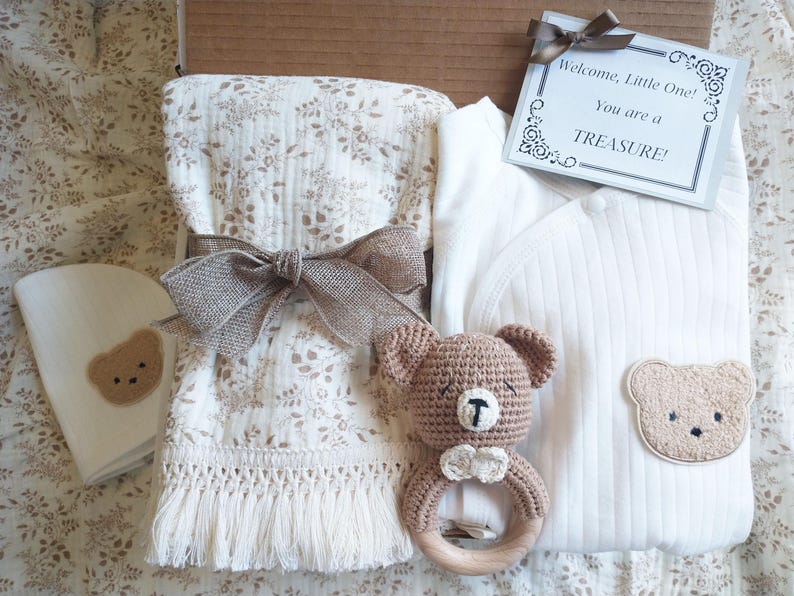Pu&ograve; includere: Un set regalo per neonati con un body bianco con un'applicazione a forma di orsetto, una coperta con stampa floreale, un sonaglio a forma di orsetto e un biglietto con la scritta "Welcome, Little One! You are a TREASURE!". Il set &egrave; legato con un nastro marrone.