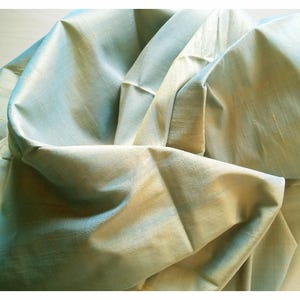 May include: Close-up of a folded fabric with a shimmering, iridescent effect. The fabric has a blend of light blue and gold tones, creating a soft, luxurious appearance. The folds and creases add depth and texture to the material.