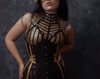 May include: A person wearing a black and gold corset with a gold and black patterned top. The person is wearing a gold mask that covers their eyes. The background is a dark blue.