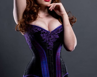 May include: A woman wearing a black and purple corset with intricate lace detailing. The corset has a boned structure and a low-cut neckline. The woman has red hair styled in a vintage updo.