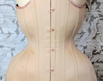 May include: A beige corset with a satin trim and rose gold metal busk closures. The corset has a curved bottom and a straight top.