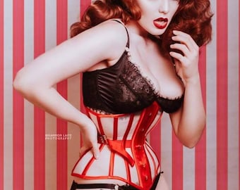 May include: A woman with red hair is wearing a white and red striped corset with a black lace bra underneath. She is standing in front of a pink and white striped background.