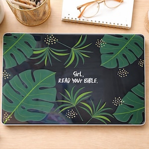Puede incluir: Funda para portátil con fondo negro y diseño de hojas tropicales verdes. El texto blanco dice "Girl, READ YOUR BIBLE". La imagen también muestra artículos de oficina y pequeñas plantas en macetas.