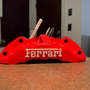 May include: A red desk organizer shaped like a Ferrari brake caliper, with the brand name in white. The organizer holds pens and pencils. The design features black accents and is placed on a brown surface.