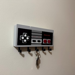 May include: A wall-mounted key holder designed as a classic Nintendo controller. The key holder is white, black, and gray with red accents, featuring the Nintendo logo and controller buttons. It has five black hooks for keys.