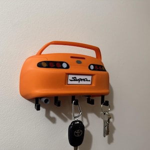 Puede incluir: Llavero naranja con forma de coche Supra montado en la pared. El llavero tiene una placa que dice "Supra". Varias llaves y un llavero de coche cuelgan del llavero.
