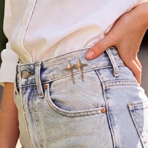 Könnte beinhalten: Eine silberfarbene Metallbrosche in Form von zwei miteinander verbundenen vierzackigen Sternen, befestigt am Bund einer hellblauen Acid-Wash-Jeans. Die Brosche hat scharfe, längliche Spitzen und eine polierte Oberfläche nahe der Vordertasche.