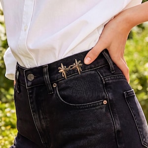 May include: A pair of metallic, star-shaped waist cincher clips in a bronze tone, attached to the waistband of black denim jeans. The clips are connected by a small metal link, featuring pointed rays and a textured, slightly melted appearance.