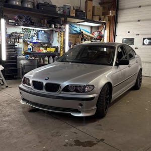 May include: Silver sedan car parked inside a garage. The car has black rims and a black front grille. The garage has tools and equipment on shelves and a closed garage door.