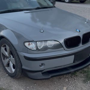 May include: A gray BMW sedan featuring a black front grille, black bumper trim, and a black front lip spoiler. The hood is equipped with two silver metal hood pins. The vehicle has clear headlight lenses and five-spoke silver alloy wheels.