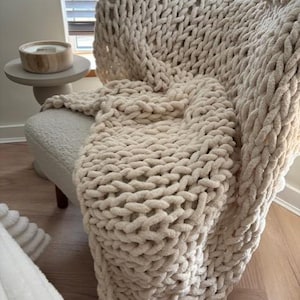 Può includere: Una coperta a maglia grossa color crema drappeggiata su una sedia. La spessa trama ad anello della coperta è visibile, creando un'estetica accogliente e invitante. La sedia è di colore chiaro e un piccolo tavolino con una candela è nelle vicinanze.