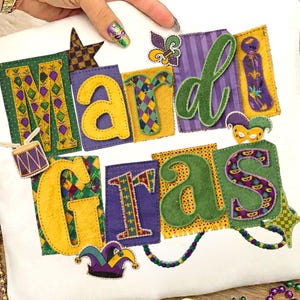 May include: White cushion featuring the words "Mardi Gras" in colourful, appliquéd letters. The letters are in various colours and patterns, with decorative elements like a drum, star, and jester's hat. The design is festive and celebratory.