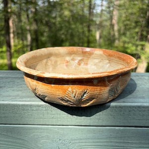 May include: A round, shallow ceramic bowl with a glazed finish in shades of orange, brown, and cream. The exterior features embossed leaf patterns pressed into the clay surface, creating a textured design around the circumference of the bowl.