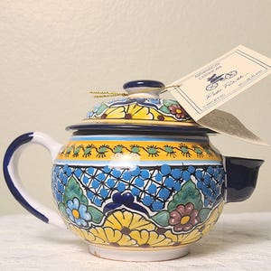 May include: Hand-painted ceramic teapot with a blue, yellow, and white floral design. The teapot features a curved handle, a spout, and a lid with a small knob. A tag with the words "Authenticity Certificate" is attached to the teapot.