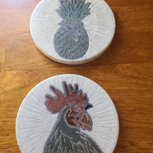 May include: Two round ceramic coasters with a cream-colored crackle glaze finish. One coaster features a dark green pineapple illustration, while the other displays a detailed profile of a rooster with a red comb and dark green plumage.