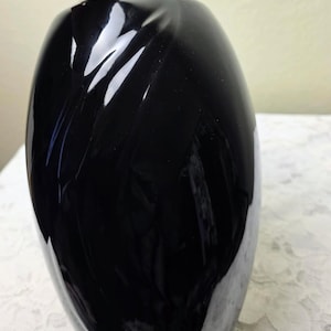 May include: A tall, glossy black vase with a rounded body and a narrow neck. The vase has a smooth, reflective surface that catches the light, creating highlights and shadows. The vase is likely made of glass or ceramic and is suitable for displaying flowers.