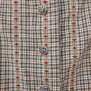 May include: A garment featuring a brown, black, and white plaid pattern with vertical stripes of green and red floral embroidery. The piece is fastened with three silver-toned heart-shaped buttons that have an embossed, intricate design.