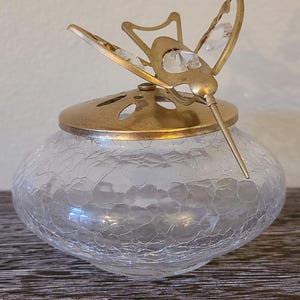 May include: A clear, crackle-glass jar with a gold-colored lid and a decorative hummingbird topper. The hummingbird has crystal accents and a long, slender beak. The jar sits on a dark wooden surface.