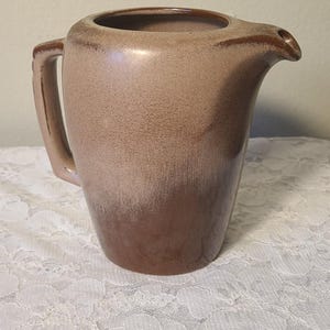 May include: A vintage ceramic pitcher with a handle and spout. The pitcher has a two-tone color scheme, with a light brown base that fades into a darker brown near the top. The pitcher is likely used for serving beverages.