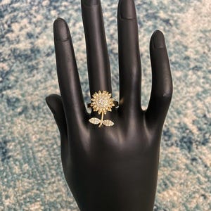 May include: A gold-colored ring in the shape of a sunflower, with a central cluster of small, clear stones and a stem with two leaves. The ring is displayed on a black hand-shaped jewelry holder.