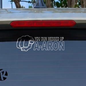 May include: A white vinyl decal applied to a car rear window featuring a graphic of a hand pointing forward with the text YOU DUN MESSED UP A-ARON in a bold sans-serif font. The design is positioned below the vehicle's red brake light.
