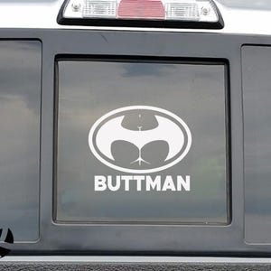 May include: White oval sticker with a humorous design. The design features a stylized image resembling a bat symbol, but with a butt shape. The word "BUTTMAN" is printed in white below the image.