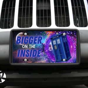 May include: A license plate with the text "BIGGER ON THE INSIDE" in blue, featuring a blue police box and a space-themed background. The plate is mounted on a vehicle.