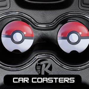 May include: Two car coasters with a red and white design, resembling a Poké Ball, are placed in a car's cup holders. The coasters have a black border and a white circle in the center. The text "CAR COASTERS" is displayed at the bottom.