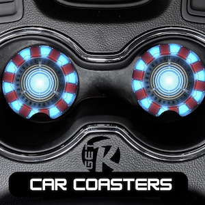 May include: Two car coasters with a glowing blue and red circular design, resembling a reactor core, sit in a car's cup holders. The coasters have a metallic silver trim. The words "CAR COASTERS" are displayed in white text on a black rectangle.