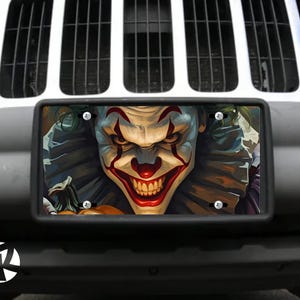 May include: A black license plate featuring a menacing clown face with red and white makeup, a wide grin, and green hair. The plate is mounted on a gray vehicle bumper, with a white grill in the background.