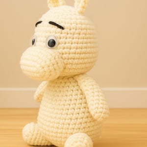 May include: A handmade, off-white crocheted hippo toy with black embroidered eyebrows and large googly eyes. The hippo has small ears and is sitting on a wooden surface. The toy is soft and plush, perfect for a child's room or nursery.