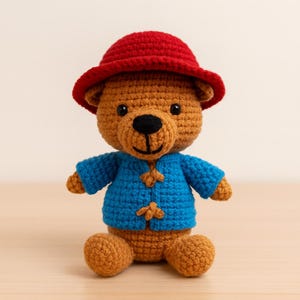 Peut inclure: Un ours en crochet fait à la main, portant un chapeau rouge et une veste bleue avec des fermetures à bascule en bois. L'ours a un corps brun, des yeux noirs et un nez noir. Le jouet est posé sur une surface brun clair.
