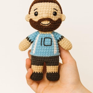 Könnte beinhalten: Handgemachte Häkelpuppe eines Mannes mit braunem Bart und Haaren, der ein hellblaues Trikot mit der Nummer 10 trägt. Die Puppe hat schwarze Shorts und beige Arme und Beine. Die Puppe wird in einer Hand gehalten.